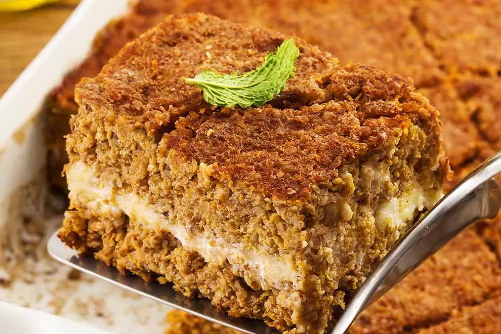 Quibe de acém com abóbora e queijo, com um pedaço dele sendo retirada, em uma travessa retangular branca, sob uma mesa de madeira.