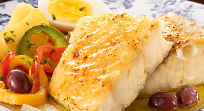 imagem destaca a receita de bacalhoada com pimentão, cebola, azeitona e ovo cozido servida em um prato redondo, de cores branco e azul, que imita um azulejo português.