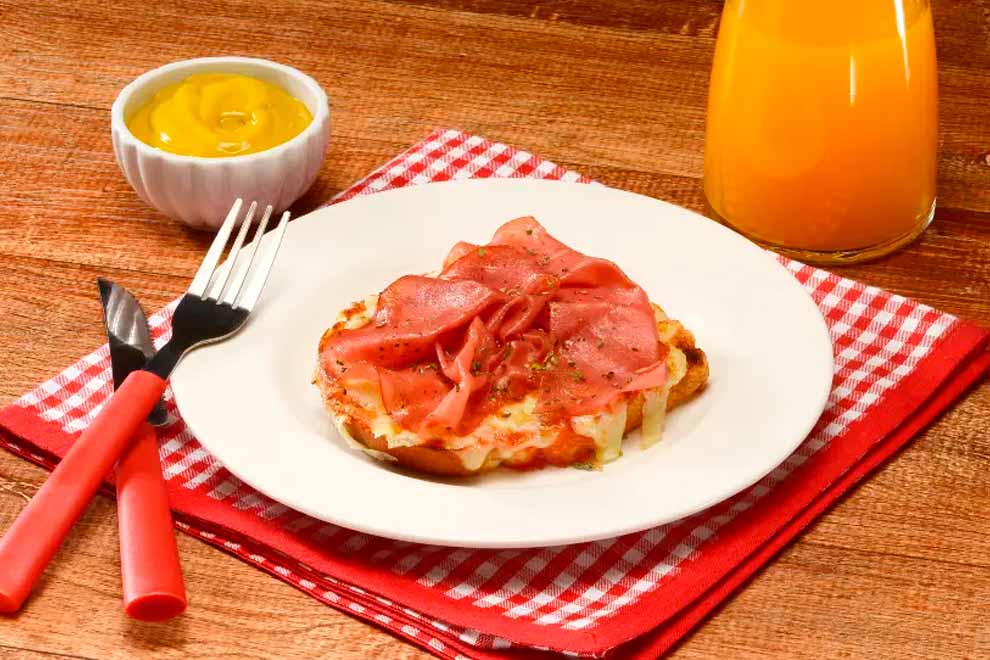 pizzinha de mortadela dentro de um prato branco de porcelana em cima de um guardanapo xadrez em vermelho e branco com talheres ao lado e em cima de uma mesa de madeira 