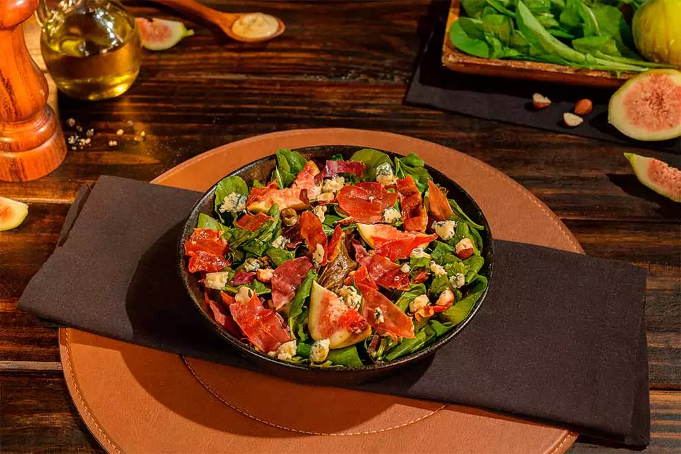 salada com presunto cru, dentro de um bowl preto em cima de um pano escuro e um jogo americano redondo em couro e cor marrom , em cima de uma mesa