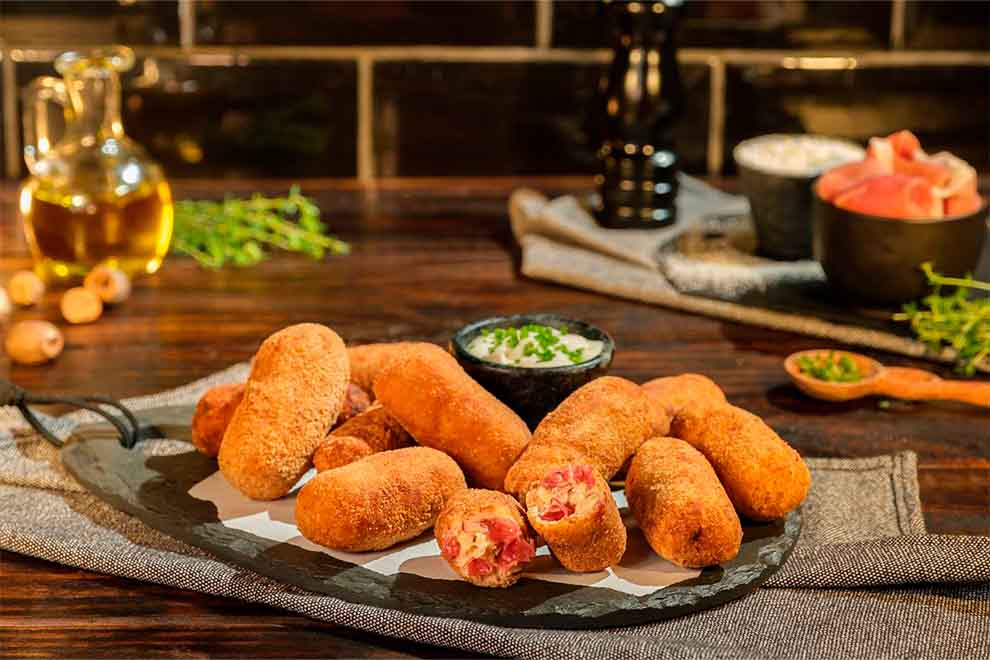 croquete de presunto jamon serrano dentro de um prato irregular de pedra e com um pequeno bowl de porcelana com molho dentro em cima de uma mesa de madeira com guardanapos cinza escuros