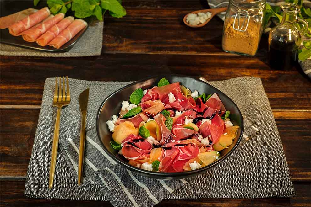 salada de presunto jamon serrano e melão dentro de um bowl preto de porcelana em cima de um pano listrado em branco e azul escuro e em cima de um jogo americano cinza escuro, em cima de uma mesa escura de madeira