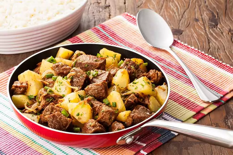 acem com batata e cebola, dentro de uma frigideira vermelha em cima de um guardanapo colorido e uma colher ao lado em cima de uma mesa