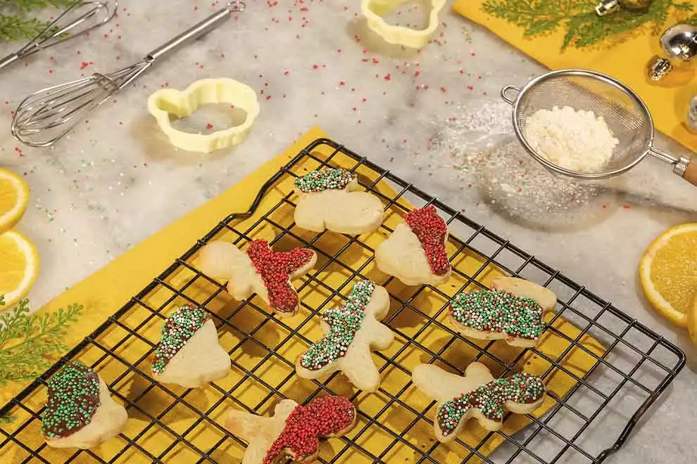 Imagem de diversos Biscoitos natalinos em cima de uma grade escura,  sob uma tábua amarela em cima de uma mesa de mármore,  com diversos ingredientes e utensílios  ao redor 