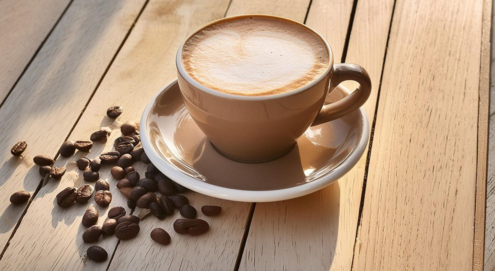 Disposição de uma xícara de cappuccino sobre um pires em uma superfície de madeira. Grãos de café torrados encontram-se ao lado. A luz natural ilumina a cena, enfatizando a categoria 