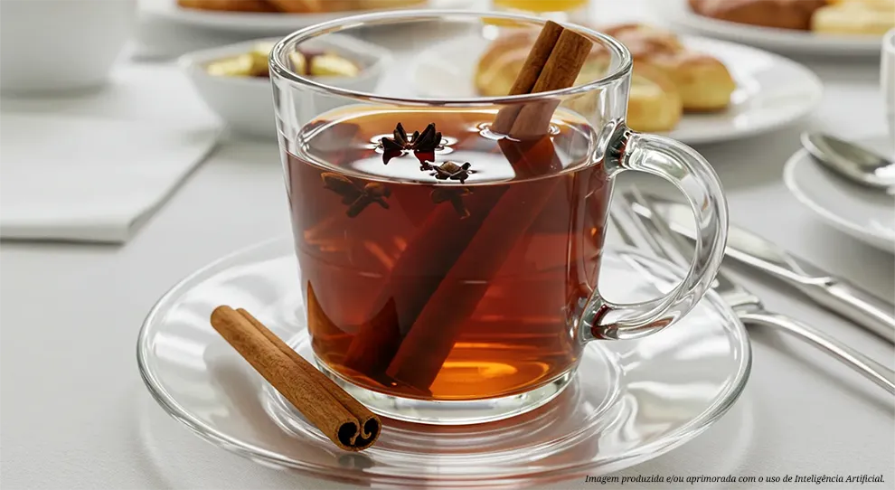 Chá de canela em um copo de vidro transparente, com pauzinhos de canela e anis estrelado submersos. O copo está sobre um pires de vidro em uma mesa. O fundo desfocado sugere um café da manhã, com 