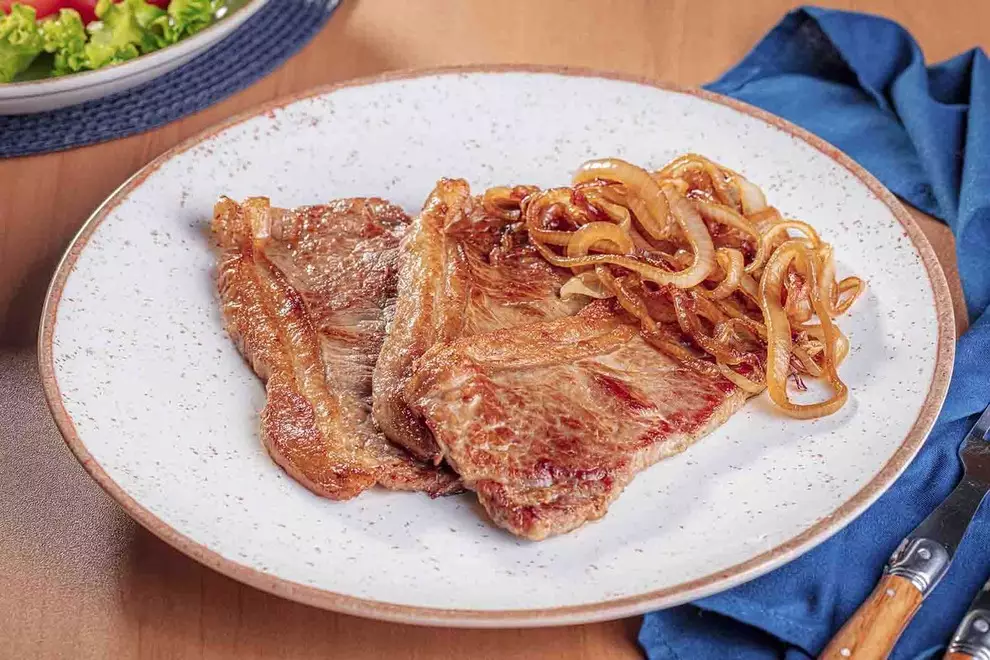 Imagem de duas unidades de Bife acebolado, em cima de um prato de cerâmica, com um par de talheres ao lado, sob uma mesa de madeira.