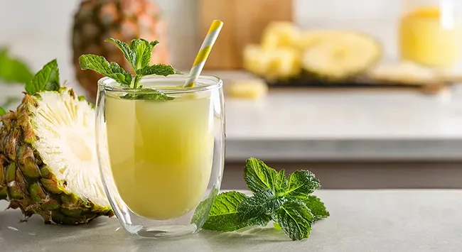 Uma bebida refrescante de abacaxi com coco em um copo estiloso, adornada com hortelã fresca e um canudo listrado, repousa sobre uma mesa. Ao lado, um abacaxi cortado ao meio revela sua polpa suculenta, com a metade superior envolta em um saquinho plástico. A mesa, com tampo de pedra, e o fundo, com cores claras, realçam a vivacidade da bebida e das frutas.