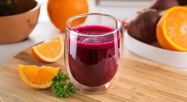 Um copo de vidro com paredes duplas está cheio de um suco vermelho vibrante, descansando sobre uma mesa de madeira rústica. A bebida, provavelmente um suco de beterraba, possui uma espuma fina e alguns respingos, indicando que foi preparada recentemente. Ao lado do copo, metades de laranjas e beterrabas criam uma composição colorida sobre a mesa. Um prato rústico com talheres e um guardanapo completa a cena, sugerindo um momento de pausa para apreciar a bebida.