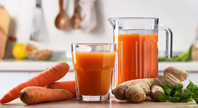 Uma foto de close-up mostra um copo cheio de suco de cenoura, um jarro cheio de suco de cenoura e três cenouras em uma mesa. Um pedaço de gengibre e um raminho de salsa estão ao lado do copo. O copo está em primeiro plano e o jarro está logo atrás dele. As cenouras estão espalhadas pela mesa. Há uma cozinha ao fundo, com armários brancos e uma bancada. A foto tem boa iluminação, com a luz vindo do lado esquerdo. A foto está em foco e as cores são vibrantes.