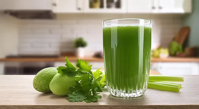 A imagem mostra um copo de suco verde fresco sobre uma superfície de madeira, com ingredientes naturais ao redor. O suco tem uma coloração vibrante e textura levemente espessa. Ao lado do copo, há limões verdes, salsinha fresca e talos de aipo, sugerindo que esses ingredientes foram usados no preparo da bebida. O fundo revela uma cozinha moderna, bem iluminada, com armários brancos e objetos levemente desfocados, criando uma atmosfera aconchegante e saudável.