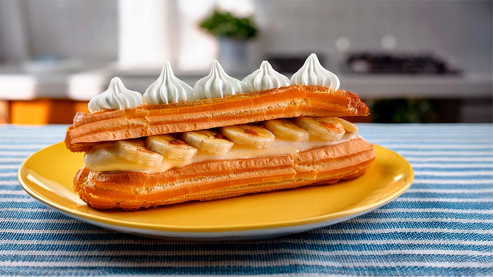 Éclair de banana com creme, merengue e massa folhada dourada, em um prato amarelo sobre uma toalha listrada.