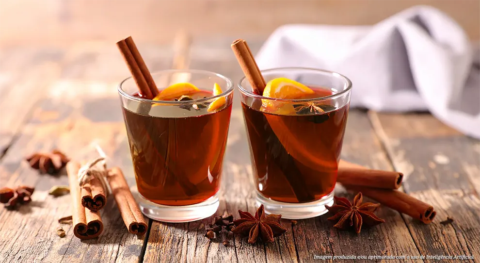 Dois copos de quentão com canela e especiarias, em uma mesa de madeira escura. Especiarias como anis estrelado, canela em pau e vagens de cardamomo estão espalhadas ao redor.
