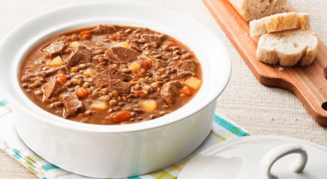 sopa feita com lentilha, músculo e mandioca. No fundo tem uma tábua de madeira com torradas.