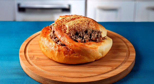 Çörek Recheado com Carne Moída servido em uma tábua de madeira sob uma mesa com toalha azul.