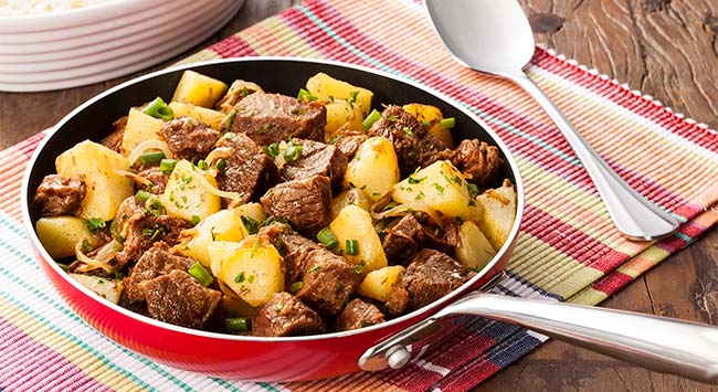Acém com cebolas e batatas servido em uma grande frigideira em uma mesa de madeira.