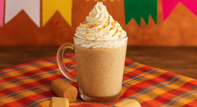 Bebida cremosa de paçoca servida em caneca de vidro com chantilly e canela por cima, em clima junino com toalha xadrez e bandeirinhas.