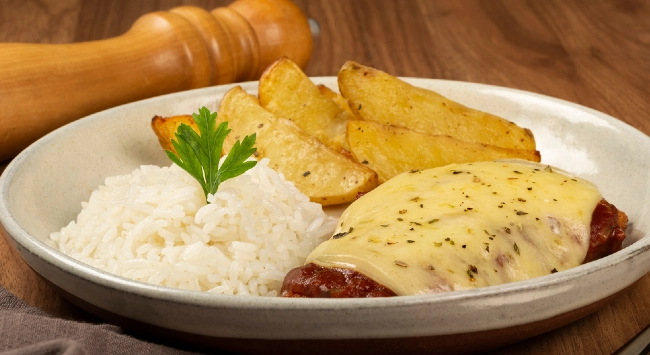 Prato com arroz branco, batatas rústicas assadas e bife à parmegiana com molho e queijo gratinado, servido em prato rústico. Feito com uma carne magra.
