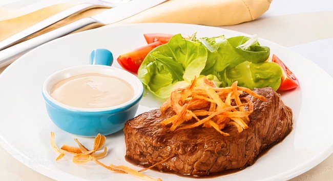 Bife de maminha com crispy de cebolinha em cima, servido com salada de alface e tomate, com um pequeno pote azul de maionese, ao lado esquerdo. Todos os elementos estão em um prato branco redondo. A foto está ampliada, bem próxima do prato.