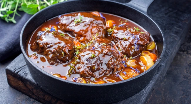 Bochechas de boi cozidas lentamente em molho escuro e brilhante, servidas em panela de ferro com legumes e ervas frescas como guarnição.