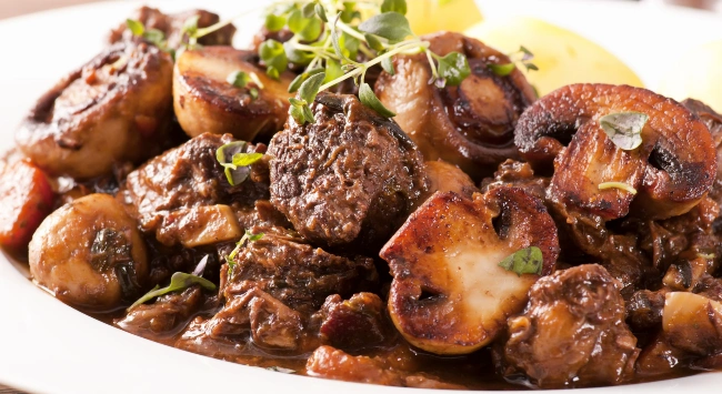 A imagem apresenta um prato de Boeuf Bourguignon, um tradicional ensopado francês. O prato é composto por pedaços de carne de músculo bovino, cozidos em um molho rico de vinho tinto, juntamente com cogumelos, cenouras e ervas frescas. Os pedaços de carne e cogumelos estão bem dourados e caramelizados, dando uma aparência suculenta e saborosa. No topo, há raminhos de tomilho fresco como guarnição. Ao fundo, podem-se ver pedaços de batata cozida, sugerindo um acompanhamento para o prato principal. A imagem captura a textura e a cor profunda do ensopado, transmitindo a impressão de um prato reconfortante e cheio de sabor.