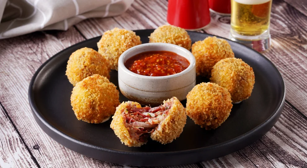 Porção de bolinhos de carne seca com parmesão e mandioca dispostos em um prato escuro. No centro do prato, há um recipiente com um molho vermelho picante. Ao fundo, um copo com cerveja.