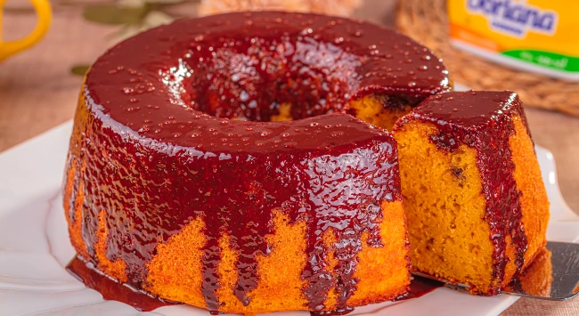 A imagem mostra um bolo de fubá em formato de anel, coberto com uma calda brilhante e espessa de goiabada derretida. O bolo, de coloração dourada e textura macia, está parcialmente cortado, revelando seu interior úmido e fofo. A calda escorre pelas laterais, criando um contraste visual atraente entre o vermelho da goiabada e o amarelo do bolo. Ao fundo, desfocada, há uma embalagem de margarina Doriana e alguns elementos decorativos, destacando o ambiente caseiro. O bolo está sobre um prato branco em uma superfície que parece ser de madeira, com iluminação quente que realça suas cores e texturas.