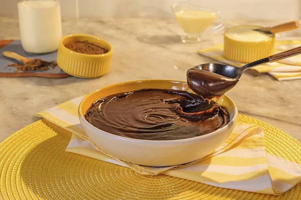 Imagem de um brigadeiro em cima de uma tigela amarela com branco, com uma colher retirando um pedaço do brigadeiro, sob um pano dobrado, sob um jogo de mesa redondo amarelo, em cima de uma mesa de mármore 