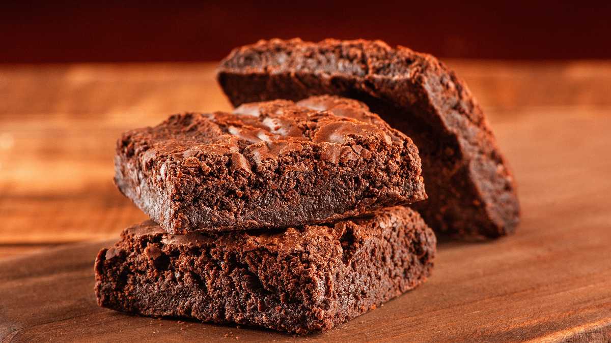 A imagem detaca 3 fatias de brownie com casquinha, empilhados em uma mesa de madeira. 