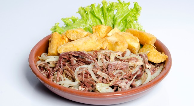 Carne desfiada acebolada servida com aipim frito em folhas de alface para decorar em um prato fundo marrom barro.