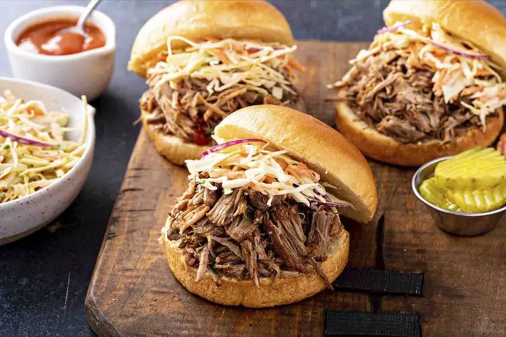 Imagem de três sanduiches de carne louca, em cima de uma tábua de madeira retangular, com alguns ingredientes ao redor, sob uma mesa escura