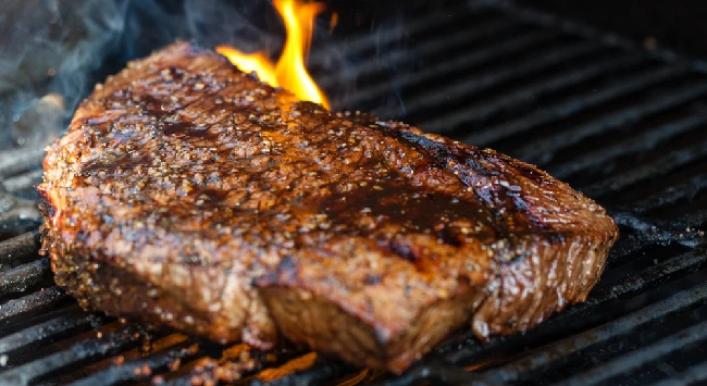 A imagem mostra um pedaço robusto de carne assando em uma grelha, com o fogo crepitando ao fundo. A carne apresenta um aspecto suculento e bem temperado, com uma crosta dourada e toques de carvão, criando um visual convidativo de churrasco.