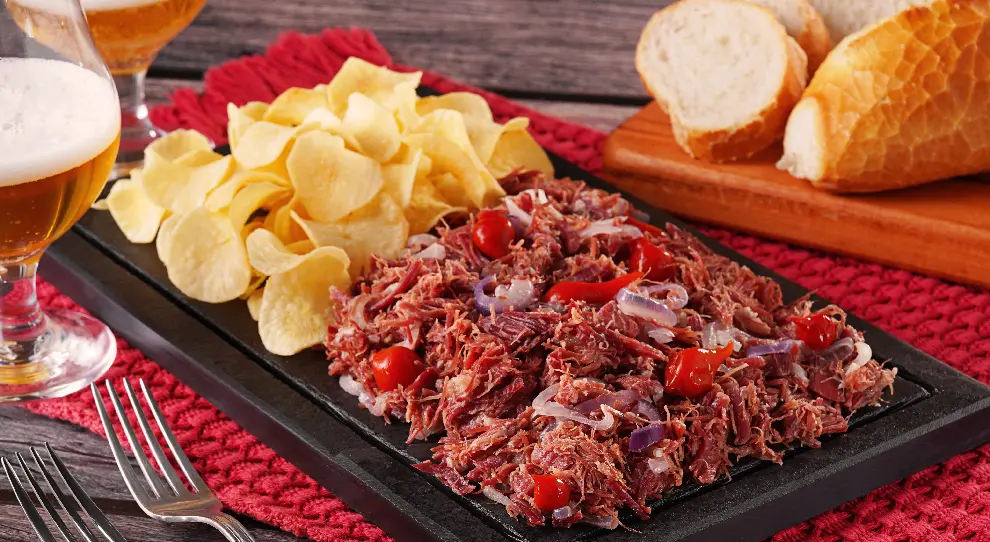 Carne seca acebolada com pimenta biquinho e chips de mandioca em uma tábua preta retangular. Ao fundo, fatias de pães e copos com cerveja.