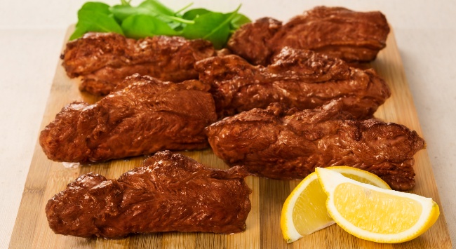 Churrasco de bananinha bovina servida em uma tábua de madeira clara com fatias de limão siciliano na frente e folhas de rúculas atrás.
