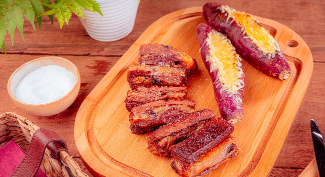 Costela assada e servida com batata doce recheada em uma tábua de madeira.