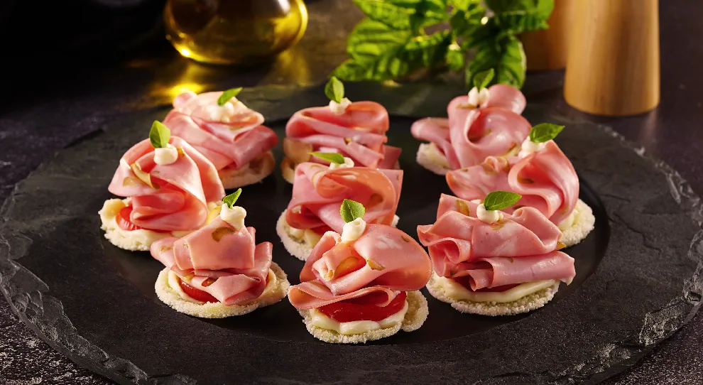 Torradinhas servidas com fatias de mortadela e queijo, em uma mesa preta.