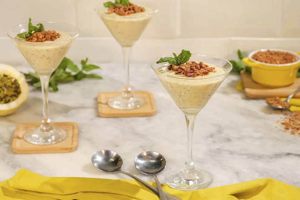 Imagem de uma taça de vidro com mousse de maracujá, com uma colher ao lado, com diversas taças com mousse de maracujá ao fundo, sob uma mesa de mármore