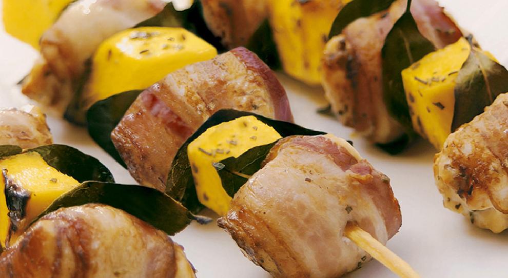 Espetinhos de medalhão de frango enrolados com bacon e cubinhos de manga enfileirados em uma mesa branca.