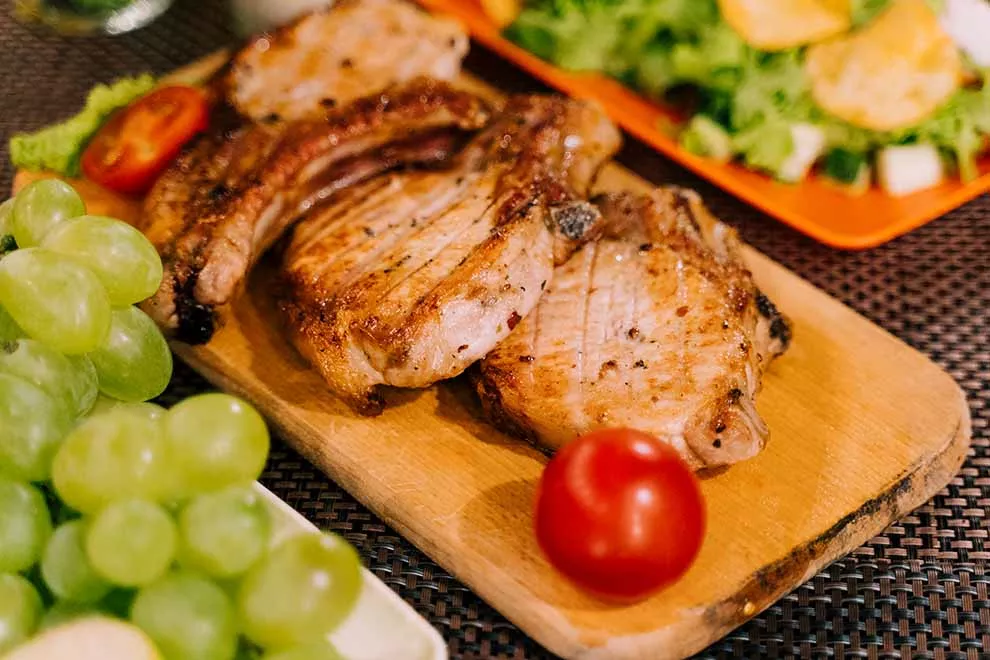alguns pedaços de carne de porco fritos em cima de uma tábua de madeira com verduras e frutas em volta em cima de uma mesa de madeira