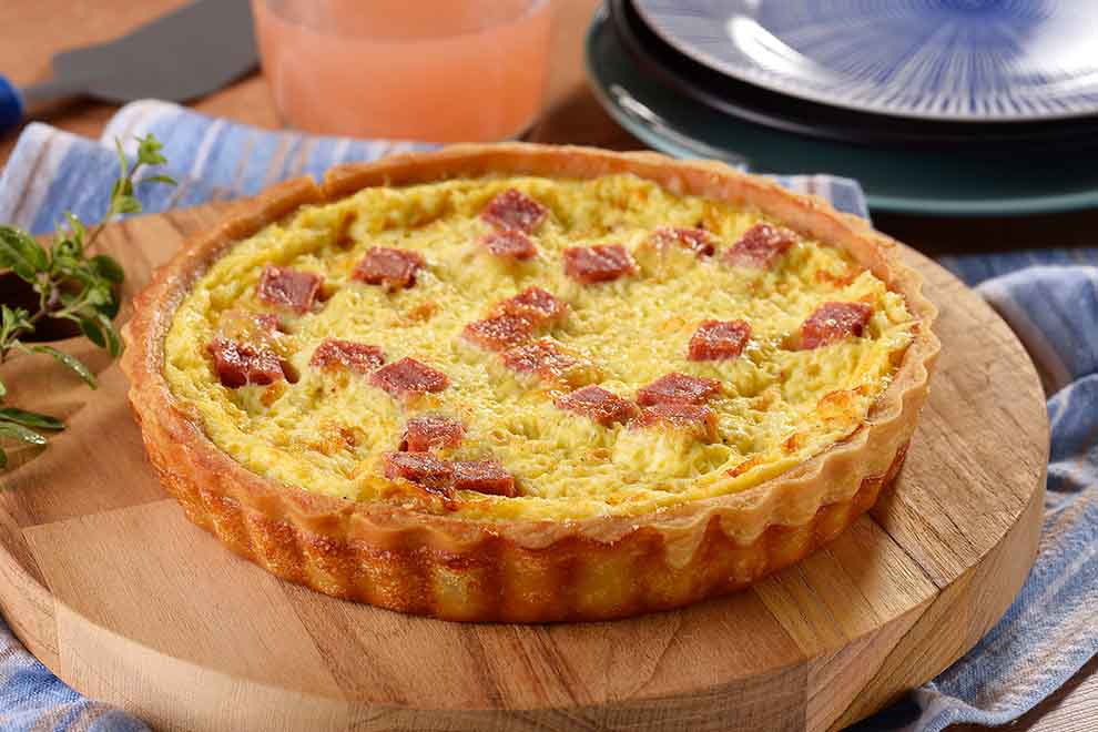 quiche de mortadela e queijo fresco em cima de uma pequena tábua de madeira redonda e em cima de um pano listrado azul 