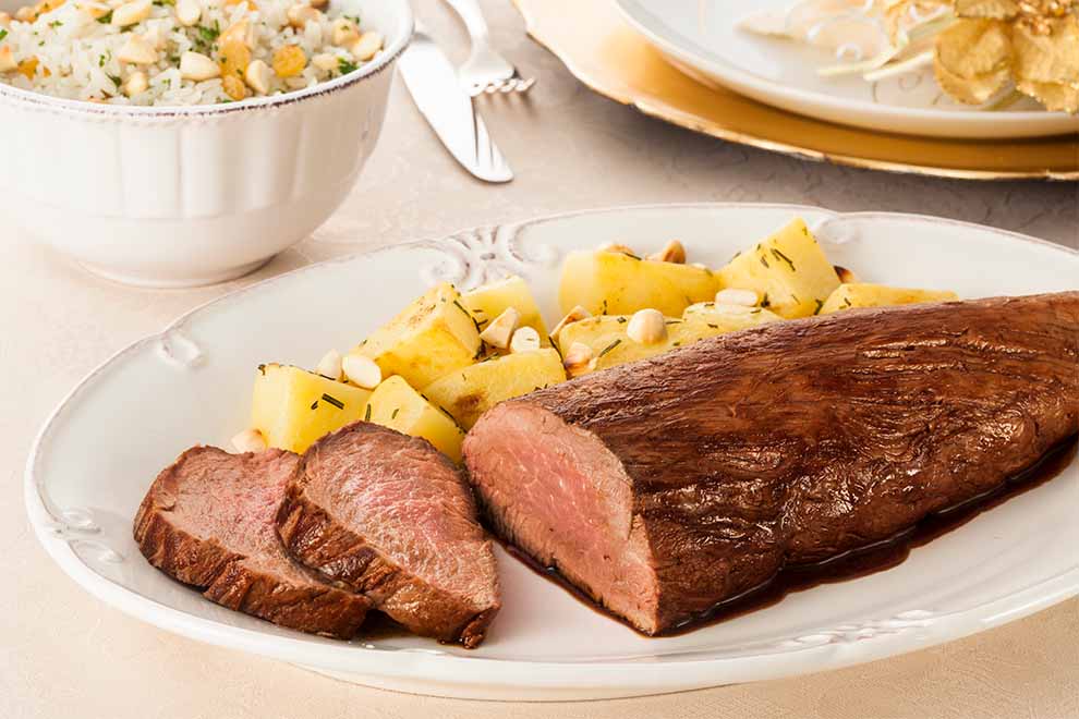filé mignon acompanhado com batata e nozes dentro de um prato oval de porcelana branco e em cima de uma mesa com uma toalha bege de mesa