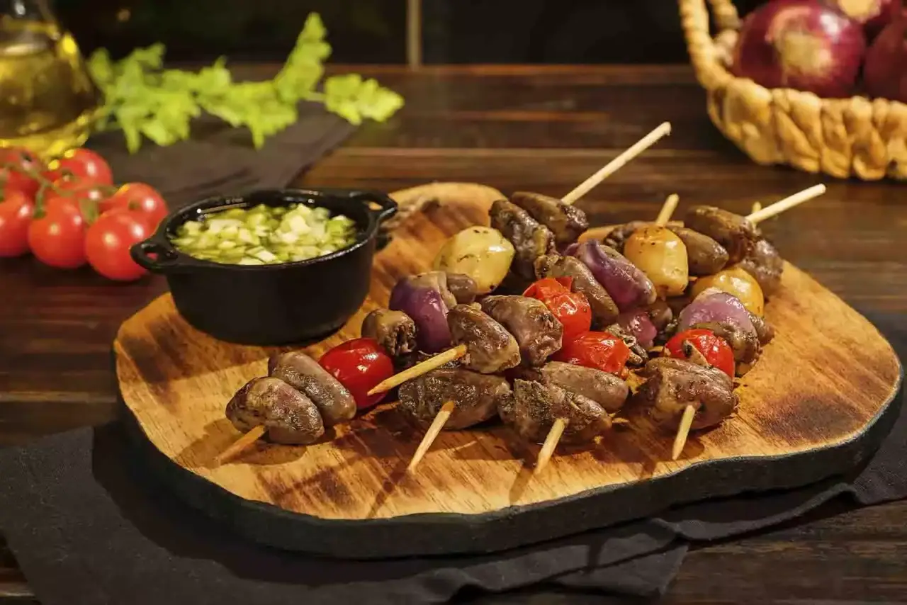 Imagem de cinco unidades de espetinho de coração, sob uma tábua de madeira com forma de troco de árvore, com uma combuca de molho ao lado, sob uma mesa de madeira com diversos ingredientes ao redor