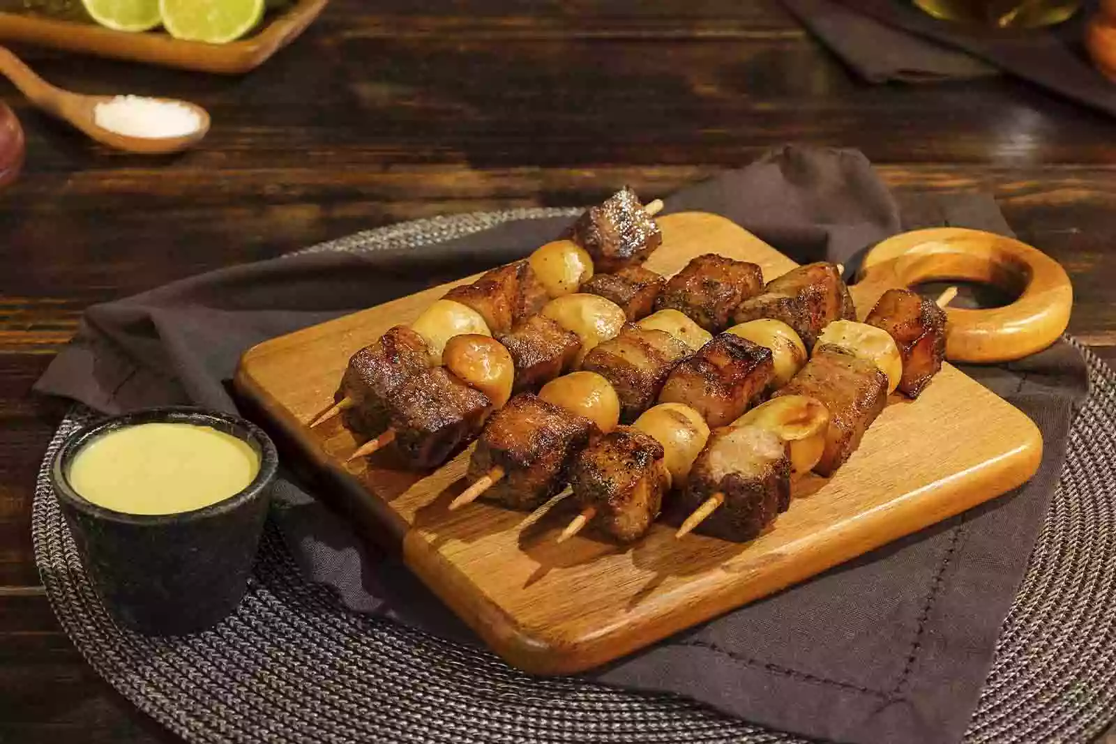 Imagem de diversos espetinhos de carne em cima de uma tábua de madeira sob um pano escuro dobrado, em cima de um jogo de mesa redondo escuro, sob uma mesa de madeira com uma combuca de molho ao lado