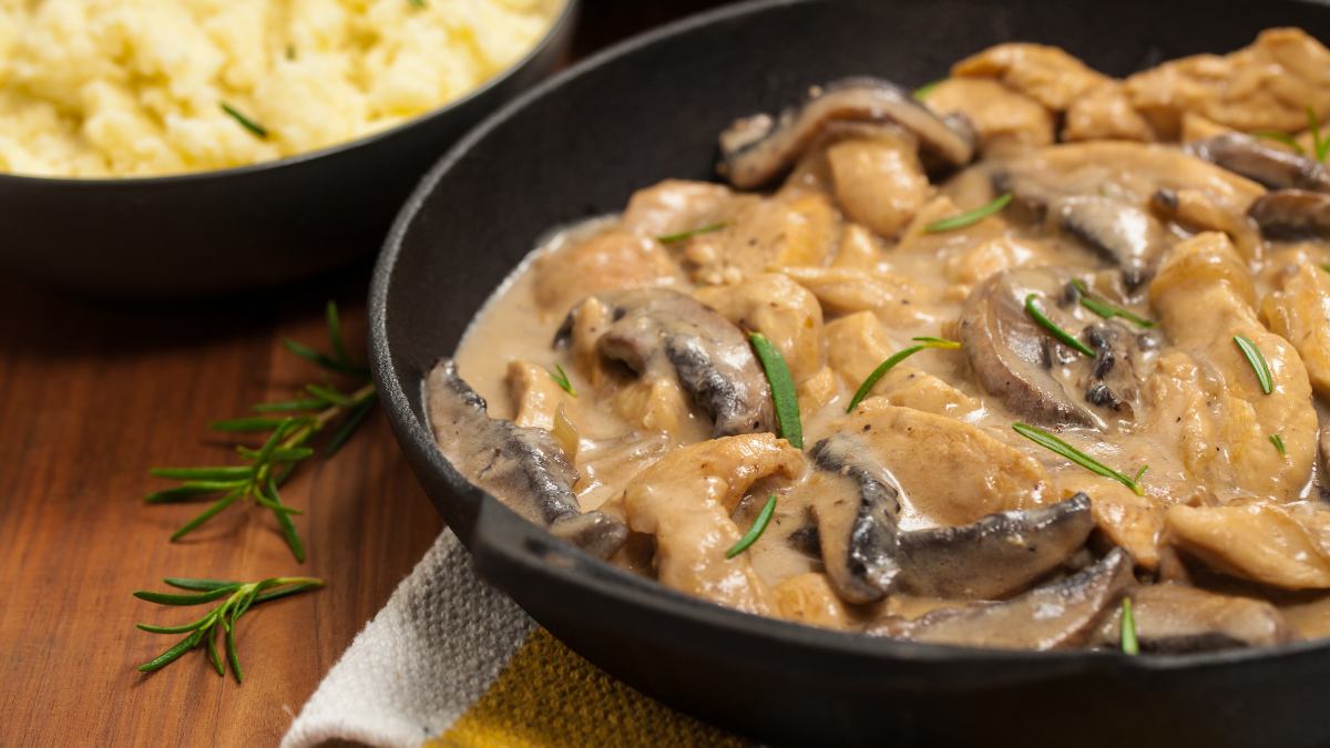 A imagem destaca o estrogonofe de frango com mix de cogumelos servido em um prato preto fundo tipo tigela. A mesa é de madeira e prato está sobre um pano listrado. No fundo há outro prato igual com farofa.