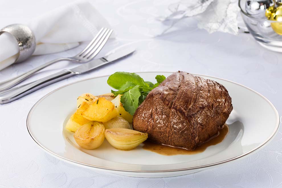 medalhão de filé mignon com mini cebolas ao lado dentro de um prato branco de porcelana e talheres ao lado e em cima de uma mesa com um pano branco 