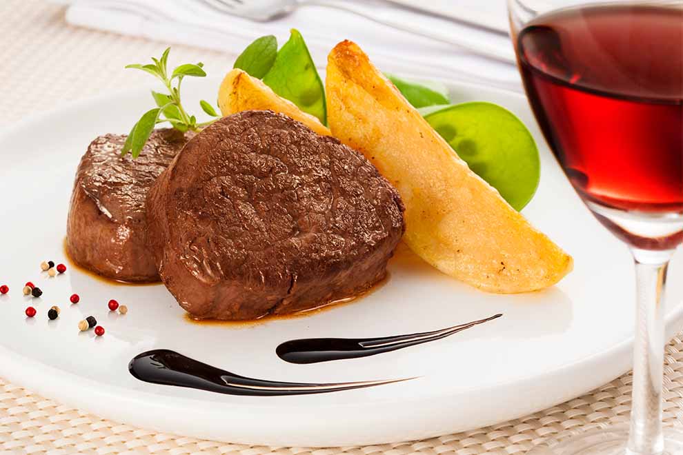 filé mignon com batata e ervilhas dentro de um prato branco de porcelana e uma taça de vinho ao lado e em cima de uma mesa com um pano branco