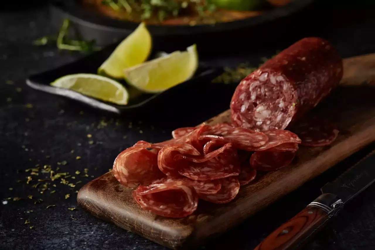 Imagem de uma peça de salame com uma parte fatiada sob uma tábua de madeira, com uma faca ao lado e fatias de limão ao fundo sob uma mesa escura 