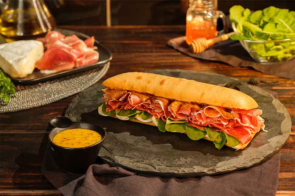 sanduíche de jamon serrano em cima de uma base em pedra e com um pequeno bowl com molho ao lado, em cima de um pano escuro e em cima de uma mesa de madeira escura