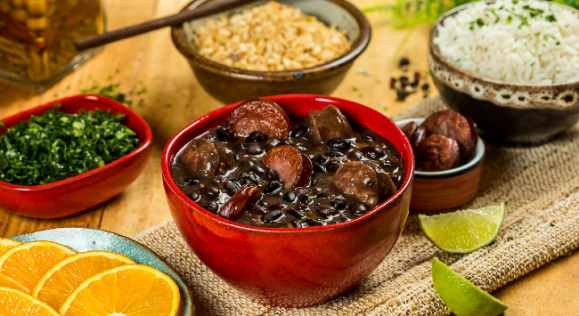 Feijoada em tigela vermelha, acompanhada de arroz, couve, farofa, fatias de laranja e limão, dispostos em uma mesa.