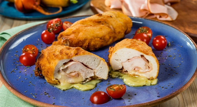 Filé de frango à cordon bleu empanado, recheado com presunto e queijo derretido, servido em prato azul com tomates cereja ao redor.